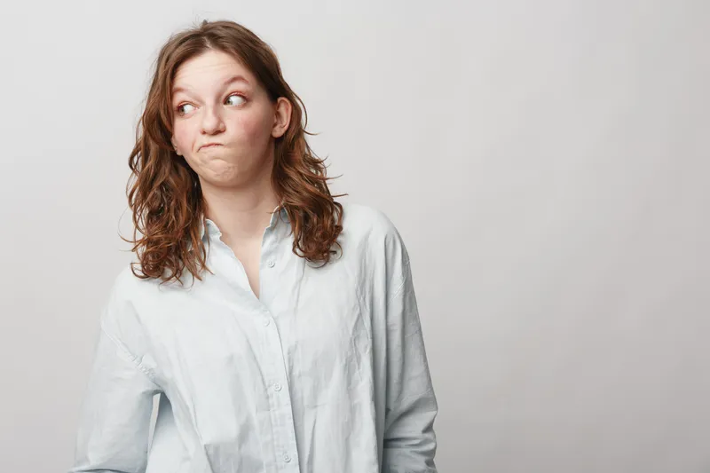 Young woman in white shirt looks sideways with skeptical expression on light background. Concept of doubt, hesitation, uncertainty, and casual lifestyle portrait.