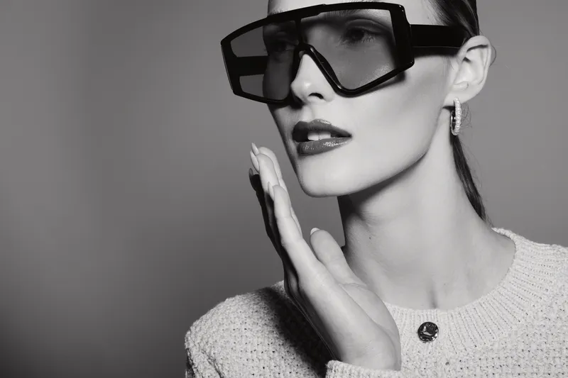 Elegant white woman with pale skin wearing oversized sunglasses and textured sweater looks sideways with hand near face in studio light. Fashion concept with confidence and style.