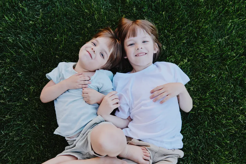 Two happy children lying on green grass, enjoying a sunny day. Their playful expressions embody joy and childhood innocence in a vibrant outdoor setting.
