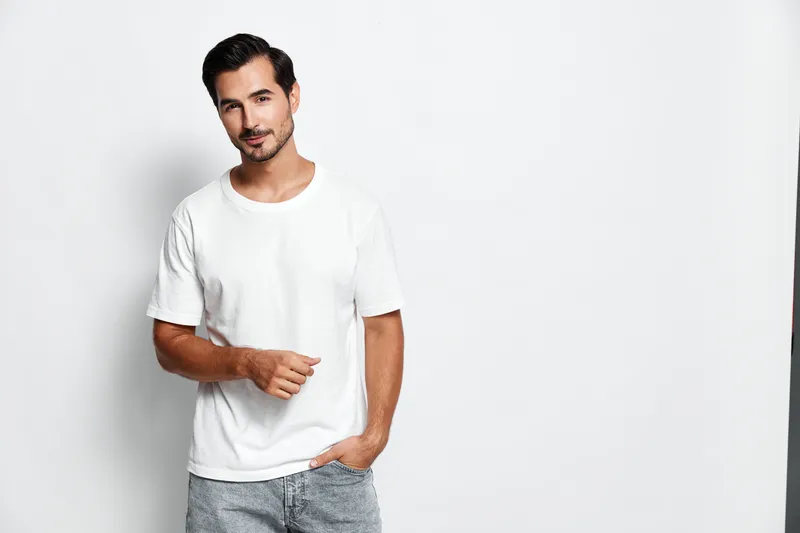 Stylish confident young man in casual white t-shirt poses against plain background with hands in pockets looking at camera with charming smile. Portrait concept