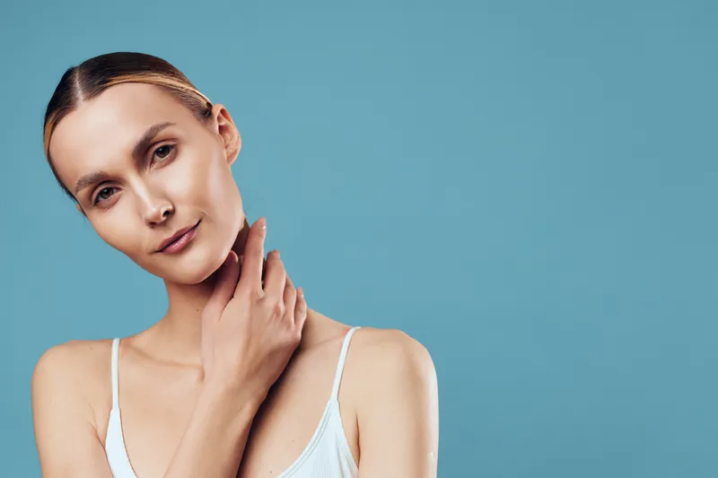 Woman with smooth face applying cream, close up portrait against blue background, soft studio lighting, calm expression, concept of skincare, beauty, wellness, self care