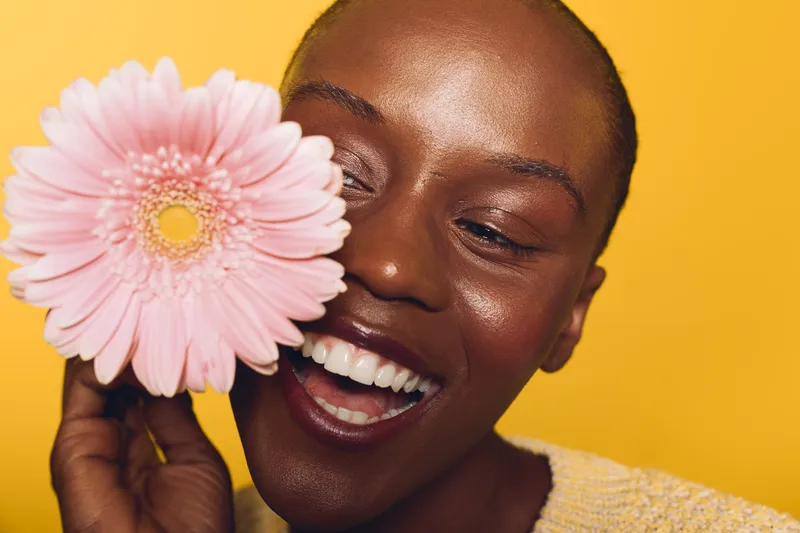 Happy African woman with dark skin smiles holding pink flower near face against yellow background. Concept of natural beauty, joy, floral, and positive mood in studio portrait.