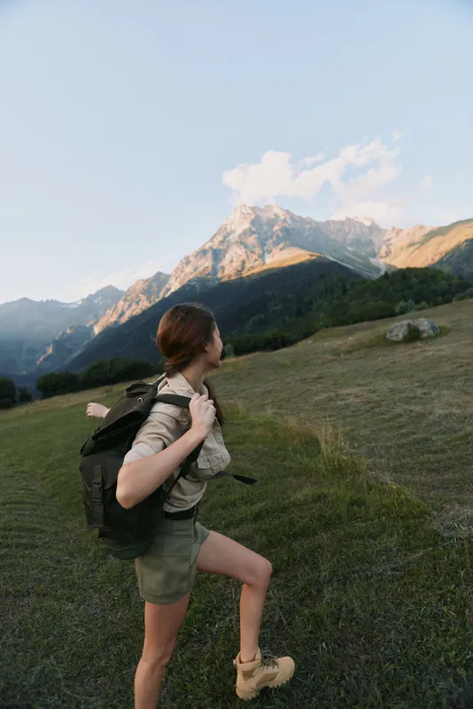 Young woman hiking with a backpack through a mountain meadow, enjoying a bright day and expansive landscape, her focused gait expresses exploration and freedom.