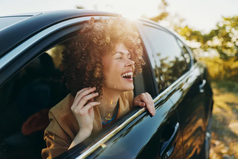 Woman with curly hair leaning from a car window, smiling as she feels the breeze. Outdoors setting with warm sunlight, relaxed lifestyle, and a carefree mood.