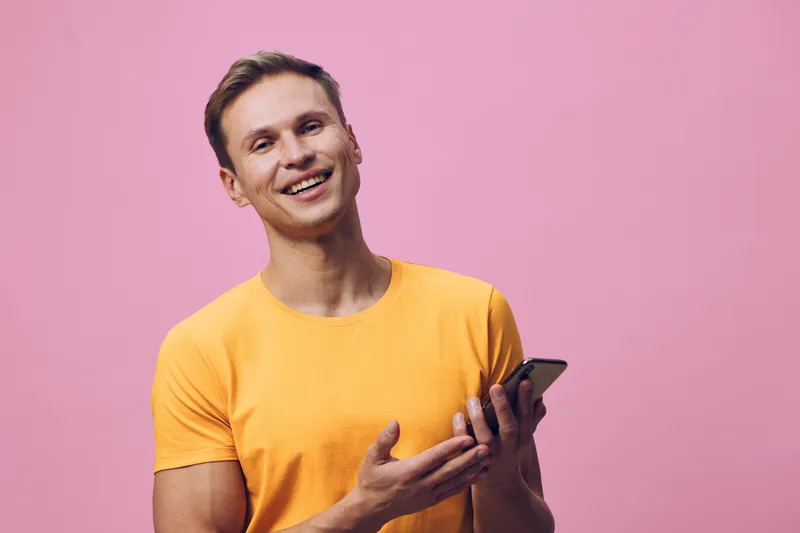 Smiling young man wearing yellow t-shirt holding smartphone on pink background, cheerful expression, casual style, bright colors, studio shot, lifestyle concept.