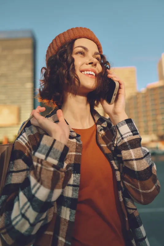 Woman on phone smile in city street wearing beanie and plaid jacket, sunlight and golden hour glow; a candid lifestyle portrait capturing authenticity, mindful living and emotional storytelling.