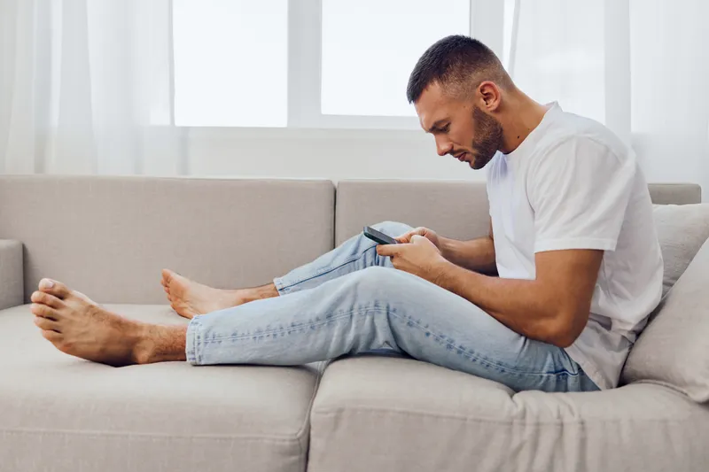 Focused man using smartphone casually seated on a cozy sofa, showcasing the modern lifestyle of digital engagement in a bright living space.
