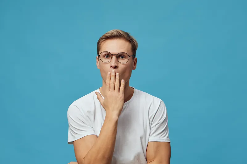 Close-up of a surprised young man with glasses wearing a white t-shirt against a blue background, covering his mouth with his hand, expressing shock or disbelief, studio portrait. People emotions conc