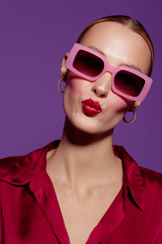 Stylish woman with pink sunglasses and red lipstick makes a kiss face on purple background. Fashion portrait with trendy accessories and vibrant colors for beauty and style concept.