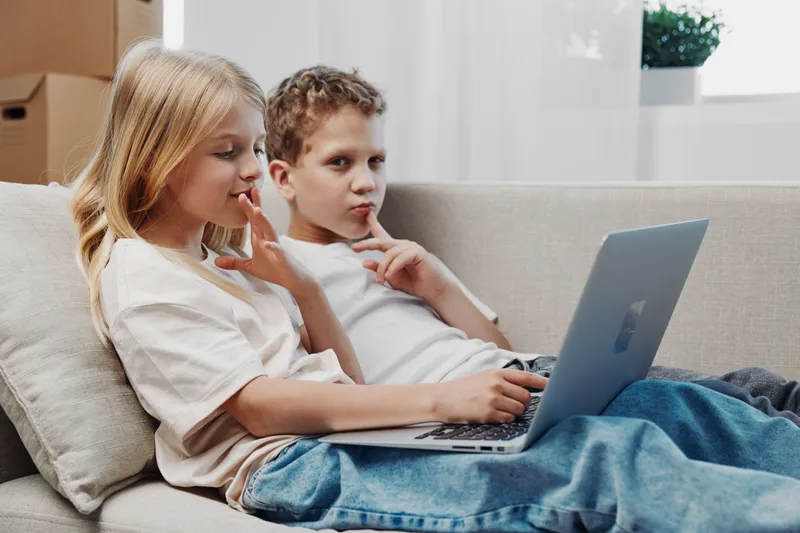 Two thoughtful children in casual clothes use laptop sitting on sofa in cozy bright room with natural light and modern interior design.