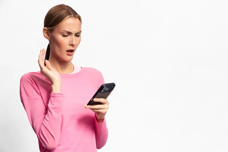 Surprised woman in pink sweater looks shocked at smartphone screen, isolated on white background. Concept of unexpected news, reaction, communication, and technology use in everyday life.