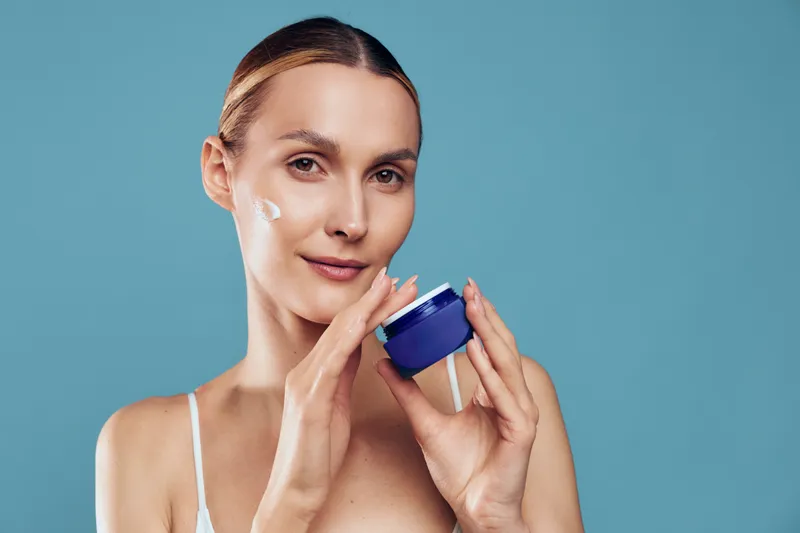 Woman applying face cream from a blue jar in a close up studio setting, calm beauty skincare concept, self care and wellness, soft studio lighting on blue background, with gentle smile