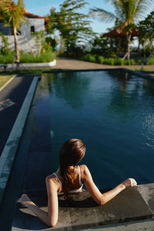 Young woman lounges by a resort pool, dipping her legs into water under warm sun, a tranquil outdoor scene with a casual summer mood and a quiet moment of rest beside palm trees.