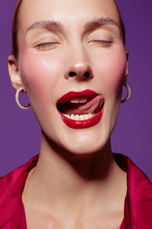 Close-up of a woman with red lipstick and closed eyes licking her lips against a purple background. Concept of beauty, makeup, sensuality, and facial expression.