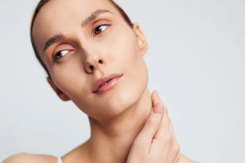 Close-up portrait of a young woman with clear skin touching her neck against a plain white background. Concept of skincare, beauty, health, and natural care with soft lighting.