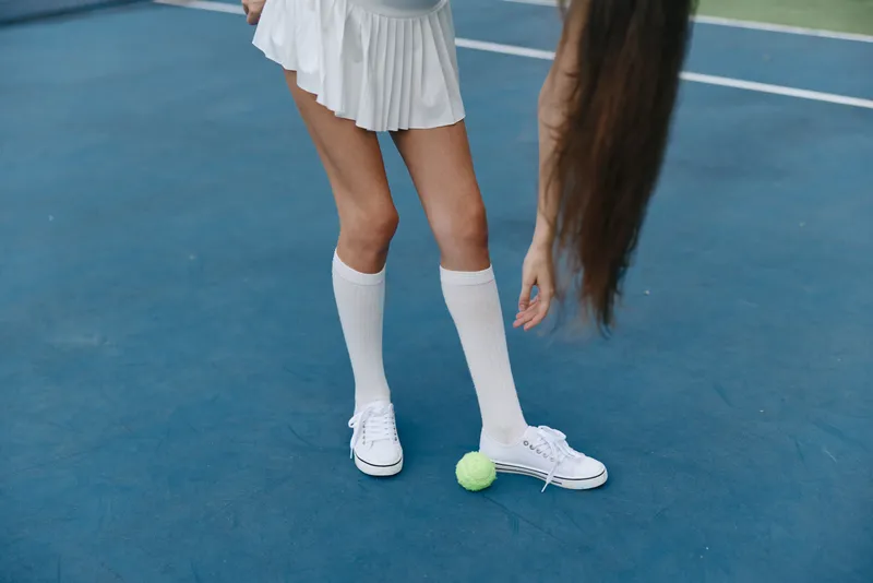 Young female athlete in white tennis attire plays on a court, showcasing white socks and sneakers, focusing on a tennis ball, vibrant blue background