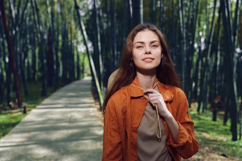 Woman walking on a forest path with a backpack and orange jacket, smiling while hiking through a bamboo nature trail. Outdoor portrait of a young traveler in casual, relaxed style enjoying sunlight