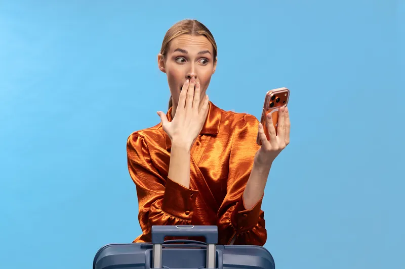 Surprised young woman in shiny orange blouse holds smartphone and covers mouth with hand, standing behind blue suitcase on blue background. Concept of travel shock and unexpected news.