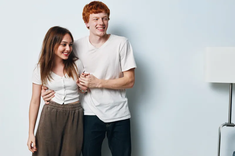 Couple, young man and woman standing close together with smiles and casual clothing against light background. Relationship, friendship and happiness with relaxed posture and natural expressions.