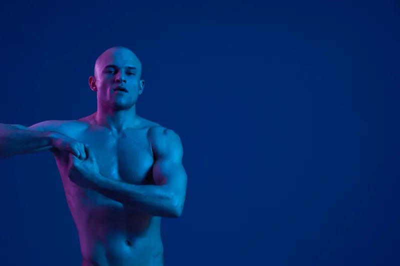 Confident man posing against a blue background showcasing strength and fitness. The vibrant colors highlight his muscular physique and artistic lighting.