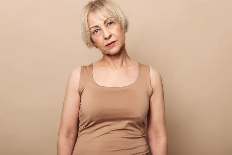 Portrait of a mature woman with short blonde hair wearing a beige tank top, looking directly at the camera against a neutral background. Concept of natural beauty and confidence.