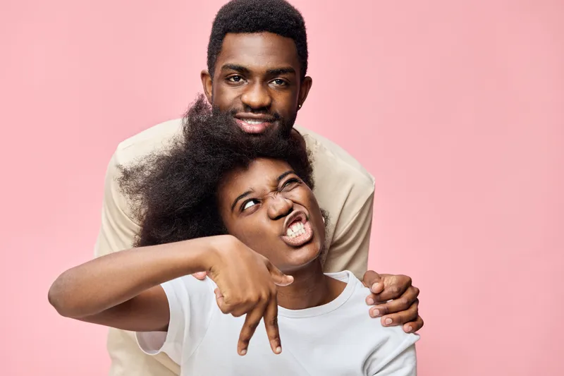 Young diverse couple smiling against a pink background, showcasing joy and affection through playful expressions Ideal for themes of love and friendship