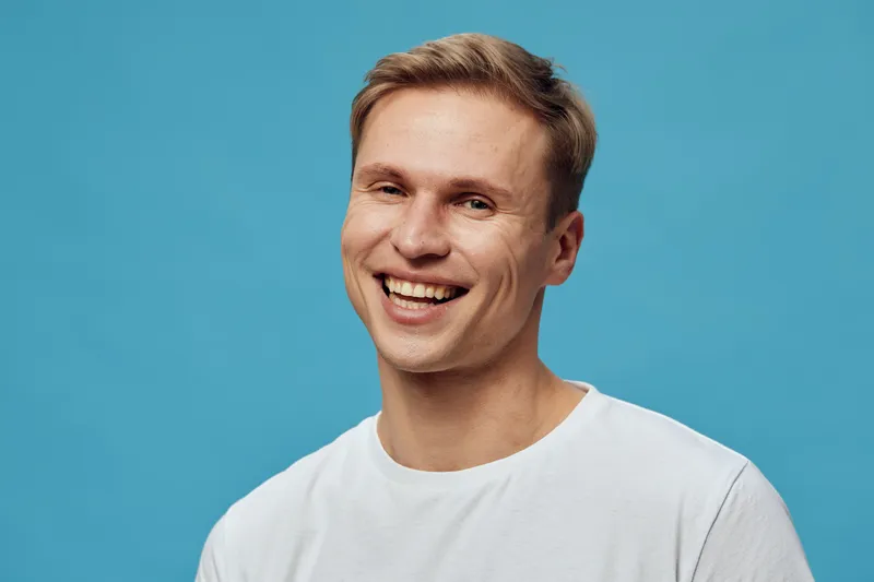 Smiling happy young caucasian man white t-shirt look camera joy mental health gadgets blue background studio portrait