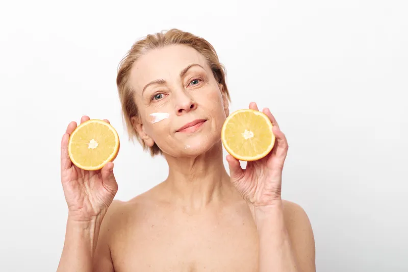 Mature woman with natural skin care holding two halved oranges and applying face cream on cheek. Concept of healthy lifestyle, beauty routine, and citrus vitamin skincare treatment.