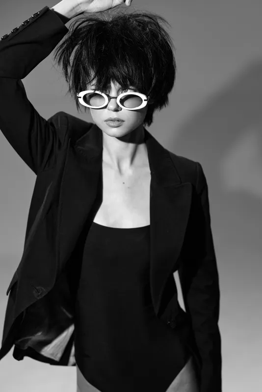 Black and white portrait of a stylish woman with sunglasses wearing a blazer and bodysuit, showing confidence and modern fashion.