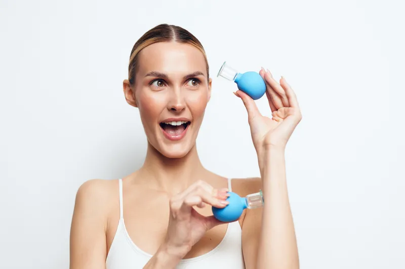 Young woman with clear skin holding two blue cosmetic bottles, smiling with open mouth, bright background, skincare routine, beauty, fresh and natural look