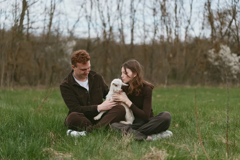 Bright couple outdoors sitting in a grassy field with a goat kid, sharing a tender moment, soft light, relaxed posture, natural colors, peaceful countryside mood