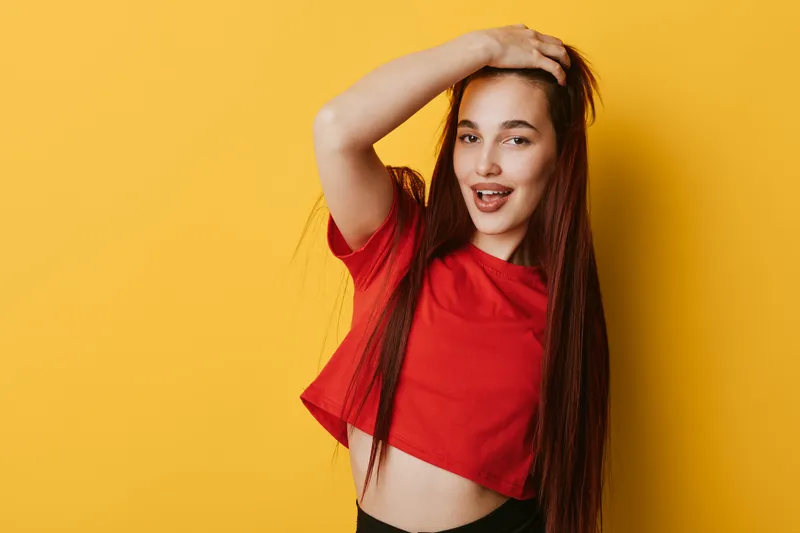 Young woman with long hair and red shirt posing with hand on head against yellow background smiling and looking at camera in studio portrait.