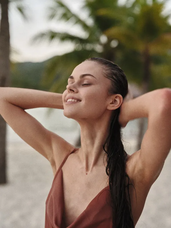 A serene portrait of a young woman on a sandy beach, eyes closed, hair wet, wearing a rust bikini, enjoying warm sunlight and a gentle breeze, radiating calm and confidence.