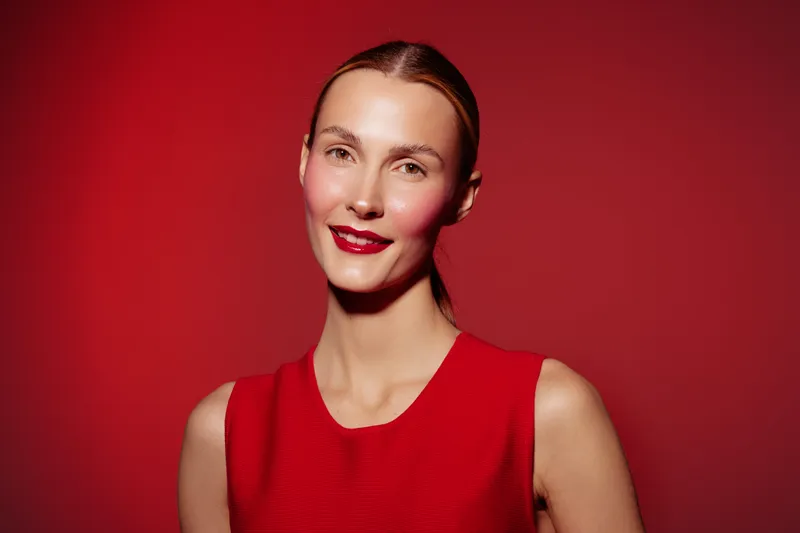 Smiling woman with red lipstick and red sleeveless dress poses against red background. Studio portrait with bright lighting highlights confident and elegant female model expression.