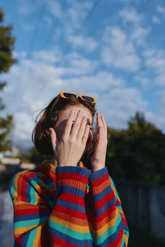 Young adult woman in a colorful striped sweater and sunglasses on her head covering face with hands, playful shy smile outdoors under blue sky in a suburban street, casual lifestyle portrait.