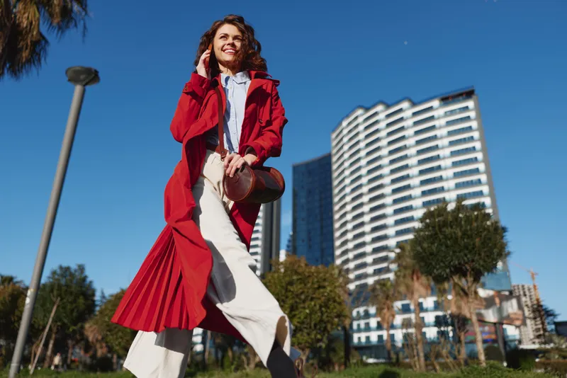 Smiling woman in red coat walking outdoors in urban park with modern buildings in background on clear sunny day. Concept of lifestyle, fashion, and city life.