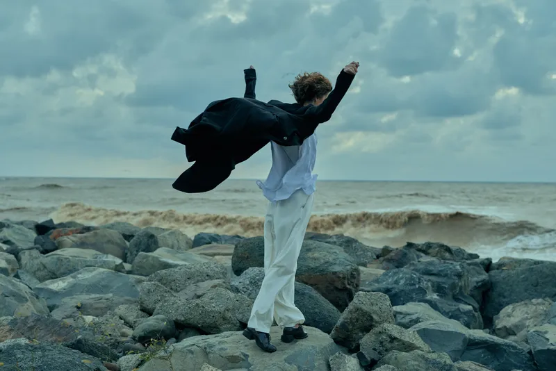 Model in white suit on rocky shoreline throws jacket with dramatic motion, strong wind, crashing waves, fashion inspired scene on a windy coast at dusk
