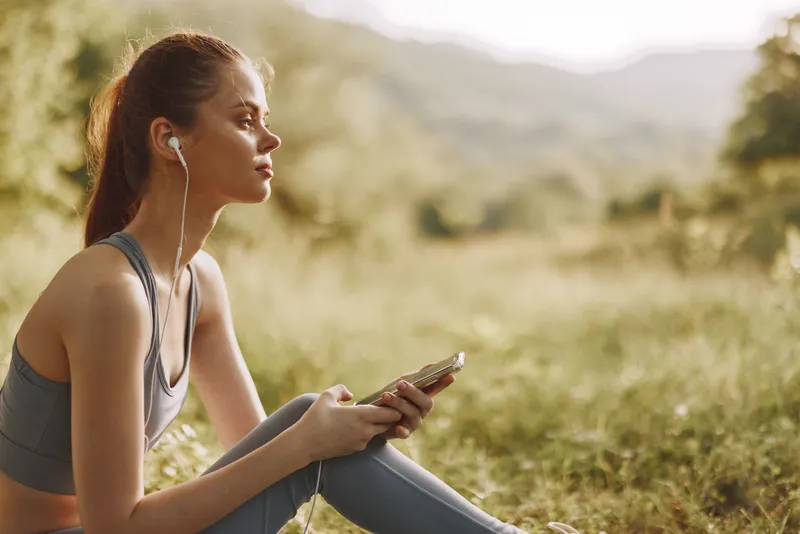 Young female athlete enjoying nature while listening to music, sitting in the grass, wearing sportswear, showcasing a relaxed and positive mood, ideal for fitness themes