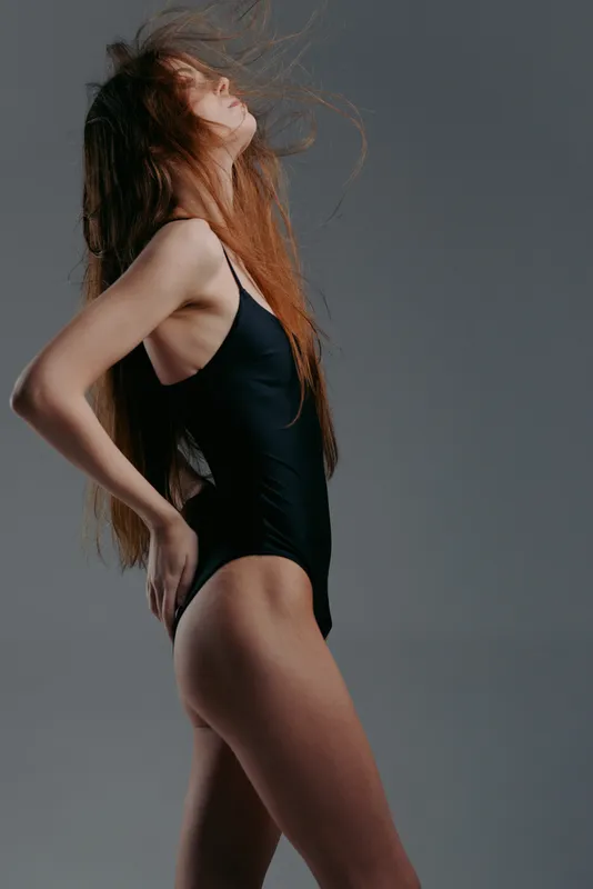 Young woman in black swimsuit posing with windblown hair on neutral background, stylish and natural beauty concept.