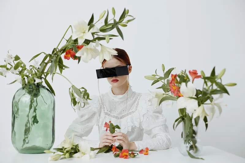A woman with red hair is sitting at a table surrounded by vibrant flowers in stylish vases, wearing a chic outfit and dark sunglasses against a minimalist white background