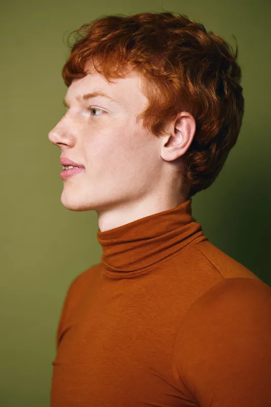 Portrait of young man with red hair wearing brown turtleneck sweater looking sideways, studio lighting with olive green background, casual fashion and natural skin tone.