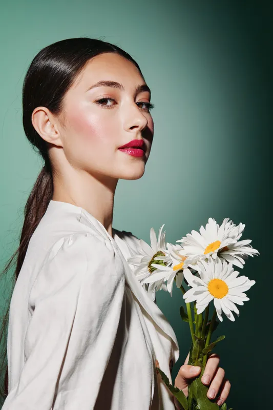 Young woman with glossy hair and natural makeup poses holding a bouquet of daisies against a green background. Concept focuses on beauty, fashion, and creative elegance.