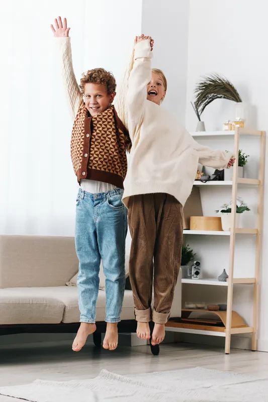 Authentic moment of playful school life as siblings jump indoors, expressing joy and curiosity in a cozy home, highlighting casual style and healthy lifestyle.