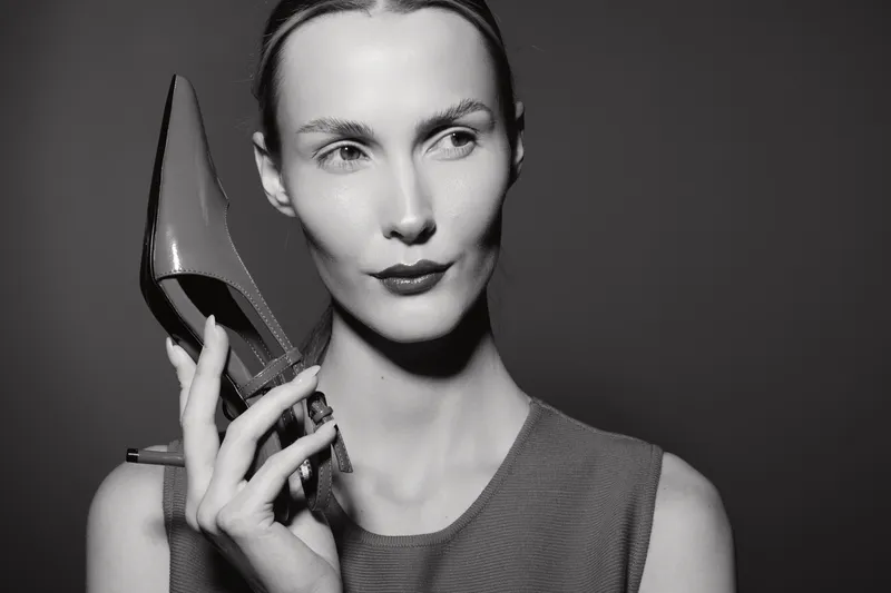 Elegant white woman with pale skin holds high heel shoe near face, wearing sleeveless top. Concept for fashion, style, footwear, and beauty in studio black and white.