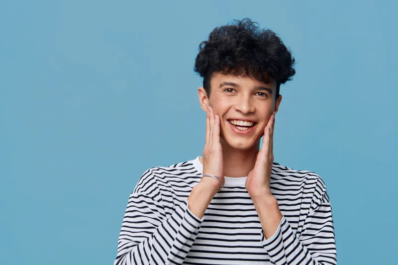 Smiling young man in striped shirt expressing joy with hands on cheeks against blue background, featuring curly hair and casual style, bright and cheerful portrait.