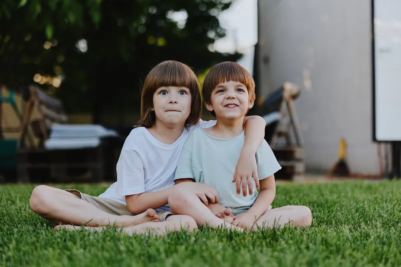 Two cute boys enjoying a sunny day in the garden, sitting on the grass and sharing a joyful moment together with bright smiles and playful expressions.