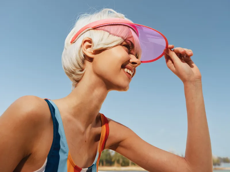 Young woman with short blonde hair wearing a pink visor smiles against a bright blue sky, capturing sunny summer style, athletic vibe, and carefree outdoor energy.