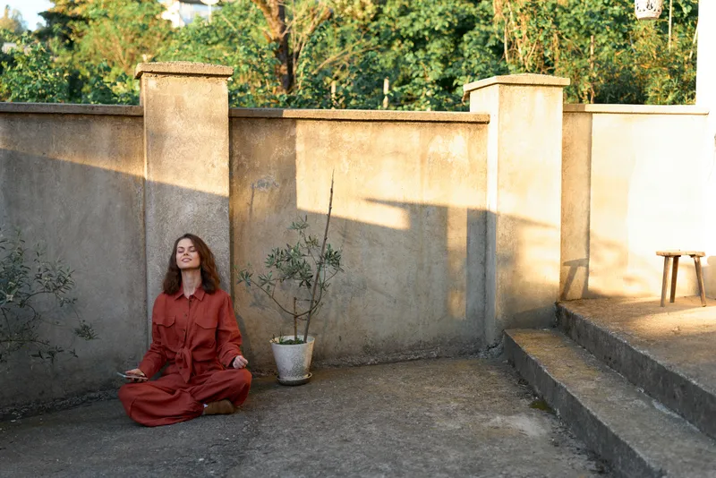 Woman in a red dress sits cross legged on the pavement beside a stone wall, a calm outdoor lifestyle portrait with natural light and serene mood