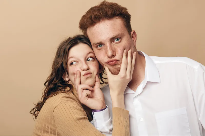 Young couple making funny faces and playful gestures, woman holding man’s face with hands. Casual clothing and beige background create a warm, lighthearted mood of friendship and fun.