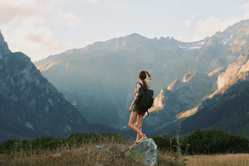 Young woman with a backpack hikes through a rugged mountain meadow, gazing toward distant peaks with a sense of adventure, independence and freedom.